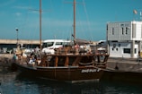 A large wooden sailing ship docked at a port with people on board. The dock is next to a modern building with a sign indicating it is a port authority. Several people are milling around the ship and dock area, with a few vehicles visible in the background.
