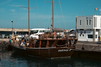 A large wooden sailing ship docked at a port with people on board. The dock is next to a modern building with a sign indicating it is a port authority. Several people are milling around the ship and dock area, with a few vehicles visible in the background.