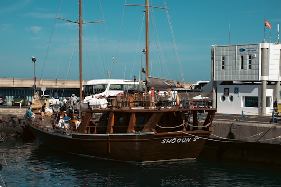 A large wooden sailing ship docked at a port with people on board. The dock is next to a modern building with a sign indicating it is a port authority. Several people are milling around the ship and dock area, with a few vehicles visible in the background.