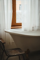 A vintage-style bathroom featuring a white, freestanding bathtub positioned under a window with white sheer curtains. A worn wooden chair is placed near the tub, and a glimpse of buildings can be seen through the window.
