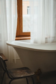 A vintage-style bathroom featuring a white, freestanding bathtub positioned under a window with white sheer curtains. A worn wooden chair is placed near the tub, and a glimpse of buildings can be seen through the window.