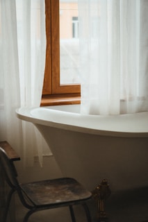 A vintage-style bathroom featuring a white, freestanding bathtub positioned under a window with white sheer curtains. A worn wooden chair is placed near the tub, and a glimpse of buildings can be seen through the window.