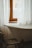 A vintage-style bathroom featuring a white, freestanding bathtub positioned under a window with white sheer curtains. A worn wooden chair is placed near the tub, and a glimpse of buildings can be seen through the window.