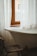 A vintage-style bathroom featuring a white, freestanding bathtub positioned under a window with white sheer curtains. A worn wooden chair is placed near the tub, and a glimpse of buildings can be seen through the window.