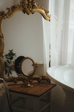 An ornate, antique-style gold mirror with elaborate detailing stands on a wooden table next to a round basin and partially covered by sheer curtains. There is a small plant visible in the background, adding a touch of greenery.