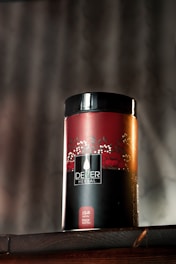 A cylindrical container with the label 'DEZER HERBAL' is placed on a wooden surface. The container features a red and black color scheme with floral designs and appears to store herbal tea. The text 'Hoor Red Tea' is visible on the bottom part of the label. A blurred background with vertical lines adds depth to the image.