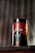 A cylindrical container with the label 'DEZER HERBAL' is placed on a wooden surface. The container features a red and black color scheme with floral designs and appears to store herbal tea. The text 'Hoor Red Tea' is visible on the bottom part of the label. A blurred background with vertical lines adds depth to the image.