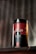 A cylindrical container with the label 'DEZER HERBAL' is placed on a wooden surface. The container features a red and black color scheme with floral designs and appears to store herbal tea. The text 'Hoor Red Tea' is visible on the bottom part of the label. A blurred background with vertical lines adds depth to the image.