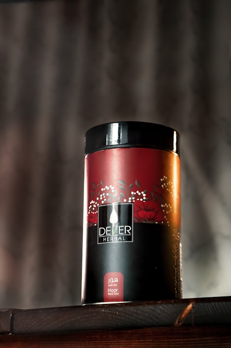 A cylindrical container with the label 'DEZER HERBAL' is placed on a wooden surface. The container features a red and black color scheme with floral designs and appears to store herbal tea. The text 'Hoor Red Tea' is visible on the bottom part of the label. A blurred background with vertical lines adds depth to the image.