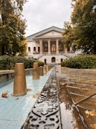 An elegant mansion featuring classical architecture with columns and large windows. It is surrounded by lush greenery and trees with autumn leaves. A decorative water feature or fountain runs along a tiled path leading towards the building, with ornamental stone cones positioned in the water.