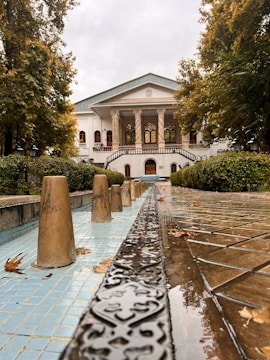 An elegant mansion featuring classical architecture with columns and large windows. It is surrounded by lush greenery and trees with autumn leaves. A decorative water feature or fountain runs along a tiled path leading towards the building, with ornamental stone cones positioned in the water.