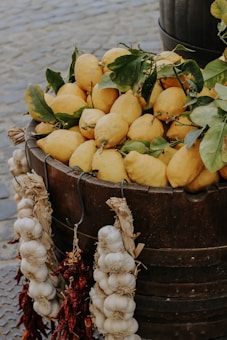 A wooden barrel is overflowing with vibrant yellow lemons, some with green leaves still attached. Strings of garlic bulbs and dried red chilies hang decoratively on the side of the barrel. The setting appears to be an outdoor market with cobblestone pavement visible in the background.