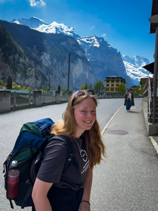 A smiling woman with a backpack standing on a scenic mountain trail during sunset.