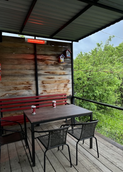 A cozy outdoor patio with a wooden deck, featuring a table with two chairs and a bench with red accents. Two coffee cups are on the table. The patio is partially enclosed with a wooden wall, adorned with small, colorful birdhouses. Lush green trees and foliage are visible beyond the patio, creating a serene natural backdrop.