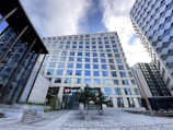 A modern cityscape featuring a tall office building with many reflective glass windows, part of a cluster of high-rise structures. The building has a prominent logo near the top and appears to house a banking institution based on the sign at the entrance. The surrounding area includes neatly arranged trees and a paved walkway, with another contemporary building visible on the right side. The sky above is partly cloudy, contributing to the urban atmosphere.