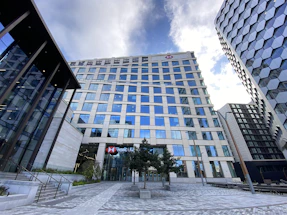 A modern cityscape featuring a tall office building with many reflective glass windows, part of a cluster of high-rise structures. The building has a prominent logo near the top and appears to house a banking institution based on the sign at the entrance. The surrounding area includes neatly arranged trees and a paved walkway, with another contemporary building visible on the right side. The sky above is partly cloudy, contributing to the urban atmosphere.