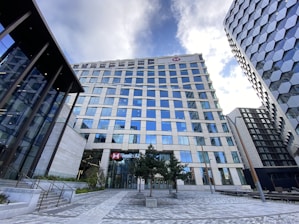 A modern cityscape featuring a tall office building with many reflective glass windows, part of a cluster of high-rise structures. The building has a prominent logo near the top and appears to house a banking institution based on the sign at the entrance. The surrounding area includes neatly arranged trees and a paved walkway, with another contemporary building visible on the right side. The sky above is partly cloudy, contributing to the urban atmosphere.