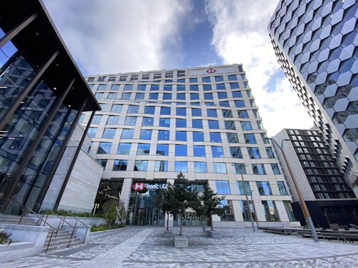 A modern cityscape featuring a tall office building with many reflective glass windows, part of a cluster of high-rise structures. The building has a prominent logo near the top and appears to house a banking institution based on the sign at the entrance. The surrounding area includes neatly arranged trees and a paved walkway, with another contemporary building visible on the right side. The sky above is partly cloudy, contributing to the urban atmosphere.