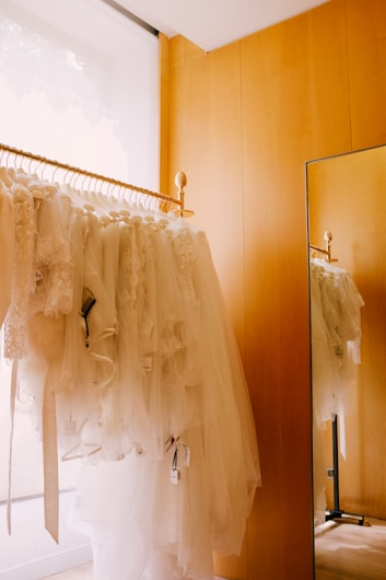 A rack filled with white bridal dresses, hanging neatly in a wooden-furnished room. There is a floor-to-ceiling mirror on the right side reflecting the dresses and part of the golden rod the gowns hang on.