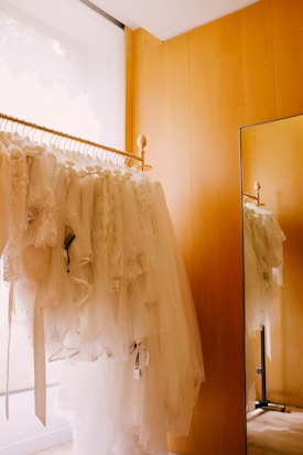 A rack filled with white bridal dresses, hanging neatly in a wooden-furnished room. There is a floor-to-ceiling mirror on the right side reflecting the dresses and part of the golden rod the gowns hang on.