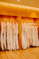 A row of white and cream-colored wedding dresses are hanging on a metal rack against a warm wooden backdrop. The floor is wooden, and the lighting is soft and warm, enhancing the elegance of the dresses.