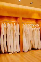 A row of white and cream-colored wedding dresses are hanging on a metal rack against a warm wooden backdrop. The floor is wooden, and the lighting is soft and warm, enhancing the elegance of the dresses.