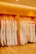 A row of white and cream-colored wedding dresses are hanging on a metal rack against a warm wooden backdrop. The floor is wooden, and the lighting is soft and warm, enhancing the elegance of the dresses.