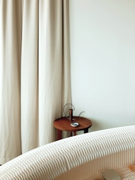 A minimalist interior scene featuring a white textured sofa in the foreground. To the side, a small wooden side table holds a metallic cylindrical object and a blue and white dish. Beige curtains and a plain white wall backdrop complete the setting.