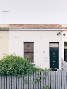 A minimalist urban facade features a simple one-story white building with two windows and a dark green door. A gray wooden fence borders the front, partially covered by lush green foliage. An outdoor light fixture is mounted beside the door, and an antenna is visible on the flat rooftop. There’s a sense of tranquility in the setting.