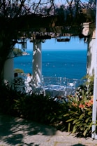 A scenic view of a terrace with elegant white furniture, surrounded by lush greenery and colorful flowers, overlooking a calm, blue ocean under a clear sky. A person is seated at one of the tables, enjoying the serene atmosphere.