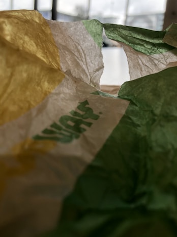 Crinkled paper with sections of yellow, white, and green, possibly part of a packaging wrapper. Blurred background indicating indoor setting with windows.