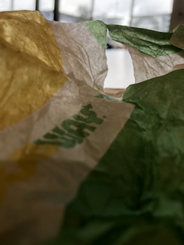 Crinkled paper with sections of yellow, white, and green, possibly part of a packaging wrapper. Blurred background indicating indoor setting with windows.