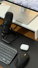 A neatly arranged workspace featuring a computer monitor with a sleek stand, a black microphone on a stand, a compact keyboard, a black ergonomic mouse, and a white charging case on a wooden surface.