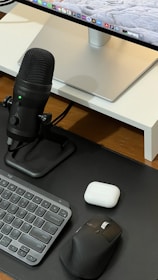 A neatly arranged workspace featuring a computer monitor with a sleek stand, a black microphone on a stand, a compact keyboard, a black ergonomic mouse, and a white charging case on a wooden surface.