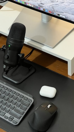 A neatly arranged workspace featuring a computer monitor with a sleek stand, a black microphone on a stand, a compact keyboard, a black ergonomic mouse, and a white charging case on a wooden surface.