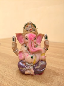 A small, intricately designed statue of Ganesha, the Hindu deity, is displayed on a wooden surface. The figure is adorned with a golden crown, pink elephantine features, and embellished with colorful jewelry and decorations. The deity's robes have a soft, purple hue and sparkle with glitter.