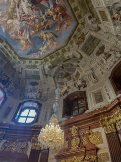 A lavishly decorated interior with an ornate chandelier hanging from the ceiling. The ceiling displays a grand fresco with vibrant colors and intricate details, surrounded by detailed architectural moldings and sculptures. The room exudes an opulent and classic style.