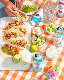 A vibrant, appetizing table setting featuring a selection of tacos with colorful toppings such as avocado, tomato, and red onion on a white platter. Various beverage cans with colorful designs are placed nearby, along with a bottle of clear liquid. A checkered orange and white tablecloth adds a festive touch. Bowls of sliced avocado, diced onions, lime wedges, and a small container of salt are arranged artistically. A person's hand is seen assembling or reaching for one of the tacos.