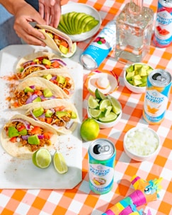A vibrant, appetizing table setting featuring a selection of tacos with colorful toppings such as avocado, tomato, and red onion on a white platter. Various beverage cans with colorful designs are placed nearby, along with a bottle of clear liquid. A checkered orange and white tablecloth adds a festive touch. Bowls of sliced avocado, diced onions, lime wedges, and a small container of salt are arranged artistically. A person's hand is seen assembling or reaching for one of the tacos.