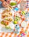 A vibrant, appetizing table setting featuring a selection of tacos with colorful toppings such as avocado, tomato, and red onion on a white platter. Various beverage cans with colorful designs are placed nearby, along with a bottle of clear liquid. A checkered orange and white tablecloth adds a festive touch. Bowls of sliced avocado, diced onions, lime wedges, and a small container of salt are arranged artistically. A person's hand is seen assembling or reaching for one of the tacos.