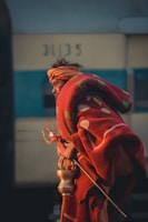 An elderly person is wearing a red and orange shawl, holding a trident and a brass lota. The person has an orange cloth wrapped around their head. The background shows a faded structure with numbers visible.