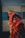 An elderly person is wearing a red and orange shawl, holding a trident and a brass lota. The person has an orange cloth wrapped around their head. The background shows a faded structure with numbers visible.