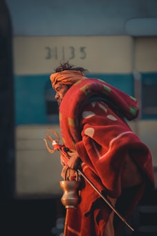 An elderly person is wearing a red and orange shawl, holding a trident and a brass lota. The person has an orange cloth wrapped around their head. The background shows a faded structure with numbers visible.