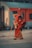 An elderly person walks along a train platform, dressed in traditional attire with a red wrap and an orange headscarf. They carry a walking stick and a metal container, possibly for water. The backdrop features a vibrant orange and gray train.