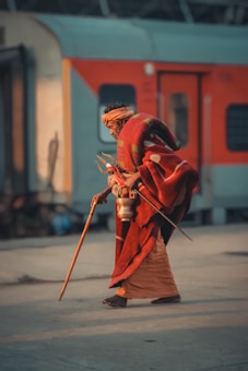 An elderly person walks along a train platform, dressed in traditional attire with a red wrap and an orange headscarf. They carry a walking stick and a metal container, possibly for water. The backdrop features a vibrant orange and gray train.