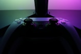 A video game controller with a sleek, modern design is positioned in front of a glowing display. The lighting casts a vibrant gradient of pink and green tones, creating a visually striking contrast against the dark backdrop.