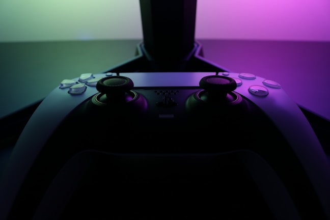 A video game controller with a sleek, modern design is positioned in front of a glowing display. The lighting casts a vibrant gradient of pink and green tones, creating a visually striking contrast against the dark backdrop.