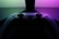 A video game controller with a sleek, modern design is positioned in front of a glowing display. The lighting casts a vibrant gradient of pink and green tones, creating a visually striking contrast against the dark backdrop.