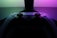 A video game controller with a sleek, modern design is positioned in front of a glowing display. The lighting casts a vibrant gradient of pink and green tones, creating a visually striking contrast against the dark backdrop.