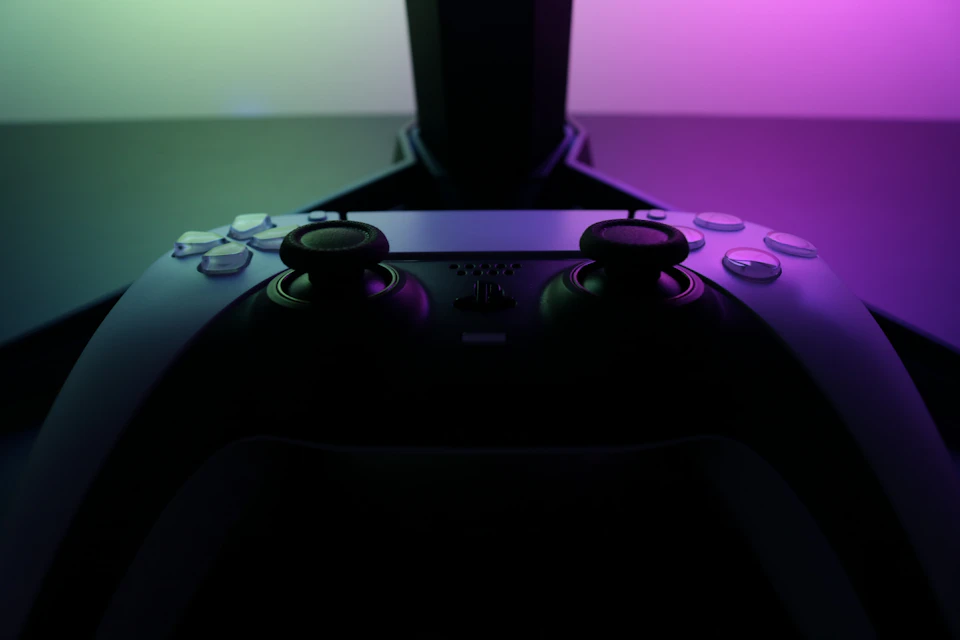 The image shows a close-up view of two black buttons on a white controller, with their tops facing upwards and slightly to the left. The buttons are located at the top center of the controller. In the background, there is a blurred purple light that appears to be coming from an unseen source, possibly a television screen or another electronic device.